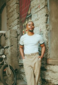 Kenny Sharp stands in a white t-shirt and khaki slacks, with hands in pockets, against a stone wall.