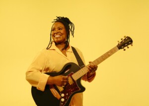 Ruthie Foster smiles while holding her guitar, wearing bright yellow in front of a bright yellow background.