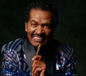 Bobby Rush smiles at the camera with his harmonica raised to his chin. He is wearing a blue sequined suit jacket, posing in front of a neutral black backdrop.